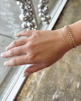 Hand wearing a gold Linked by Lustora permanent bracelet with a blurred decorative background